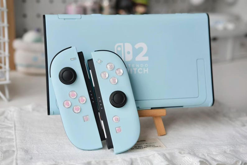 May include: A light blue Nintendo Switch console and two matching Joy-Con controllers. The console features the white Nintendo Switch logo. The controllers have black joysticks and buttons decorated with small pink heart icons on the face buttons.