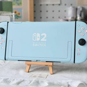 May include: A light blue Nintendo Switch console featuring a custom shell with a white Nintendo Switch logo and the number 2 printed on the back. The handheld gaming device includes black joysticks and white buttons, resting on a small wooden easel.