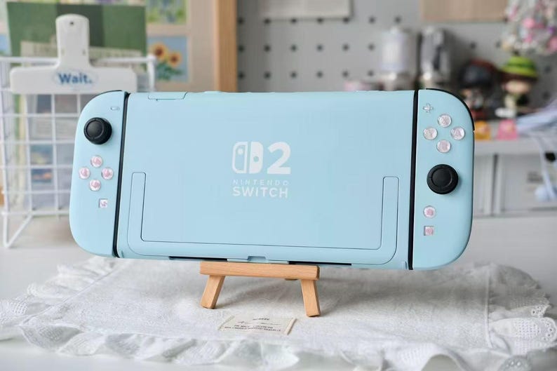 May include: A light blue Nintendo Switch console featuring a custom shell with a white Nintendo Switch logo and the number 2 printed on the back. The handheld gaming device includes black joysticks and white buttons, resting on a small wooden easel.
