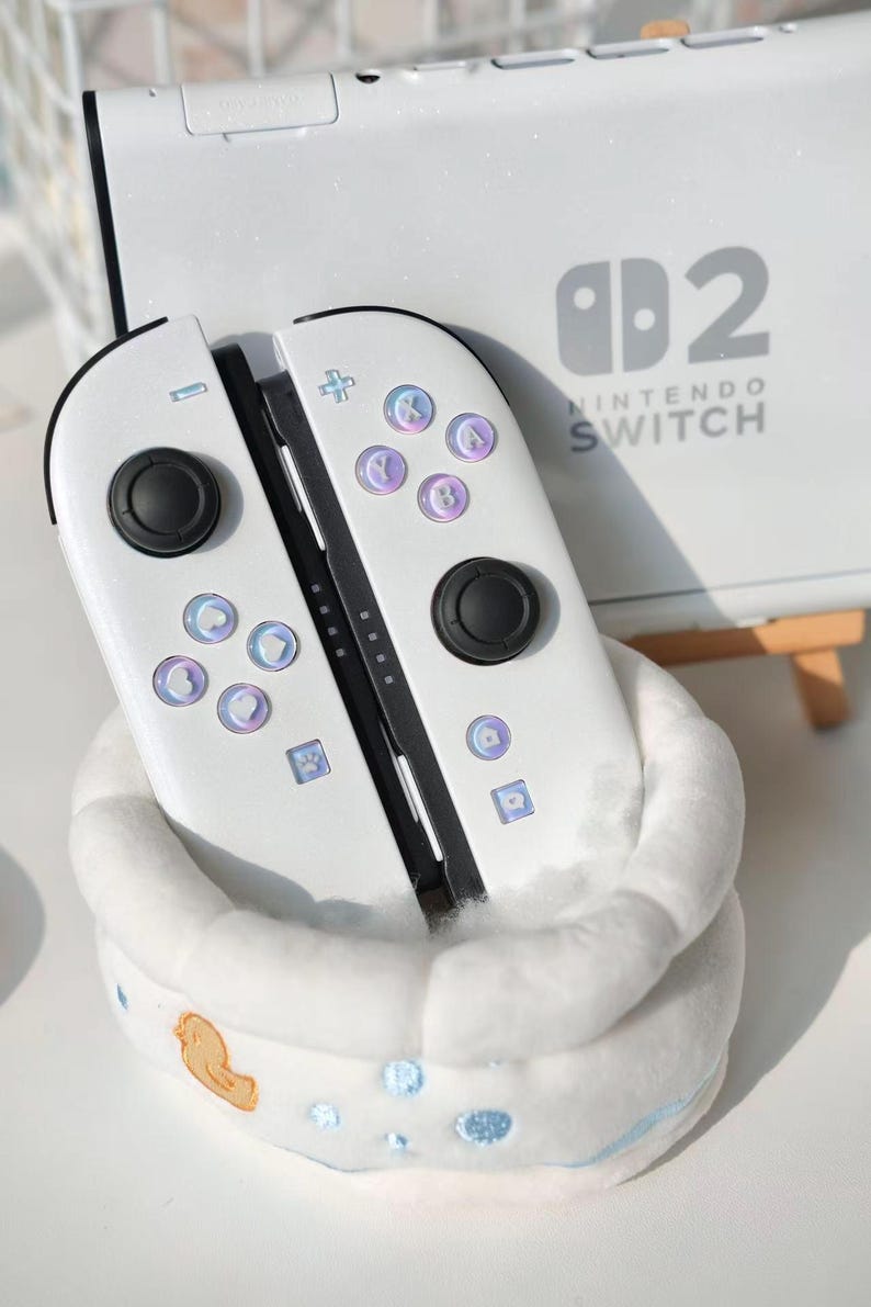 May include: White Nintendo Switch Joy-Con controllers with iridescent buttons and black joysticks. The controllers are in a white plush holder. The Nintendo Switch console is visible in the background.