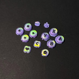 Könnte beinhalten: Eine Sammlung von irisierenden lila Gamecontroller-Tasten mit verschiedenen Symbolen und Buchstaben, darunter A, B, X, Y, ein Herz, ein Haus und Richtungspfeile. Die Tasten sind auf einer dunklen Oberfläche angeordnet.