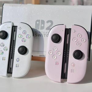 May include: Two Nintendo Switch Joy-Con controllers, one white and one pink, with black buttons and iridescent accents. A white Nintendo Switch console is in the background. The controllers have a glittery finish.