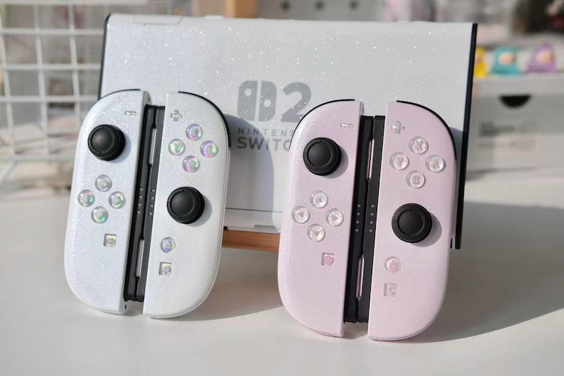 May include: Two Nintendo Switch Joy-Con controllers, one white and one pink, with black buttons and iridescent accents. A white Nintendo Switch console is in the background. The controllers have a glittery finish.