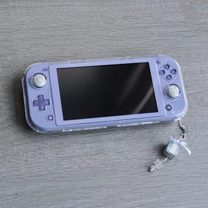 May include: A purple Nintendo Switch Lite handheld gaming console with a clear protective case. The console has a white button on the left side and a white button on the right side. A small white gift box charm is attached to the console.