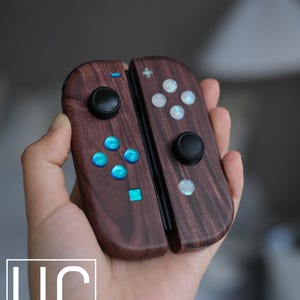 以下が含まれることがあります： ダークウッド仕上げのカスタムニンテンドースイッチコントローラー。コントローラーは青と白のボタンと、前面に黒と白のロゴが付いています。ロゴには「UC Usman's Consoles」と表示されています。