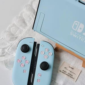 May include: A light blue Nintendo Switch console and a pair of matching Joy-Con controllers with black joysticks and light pink button accents. A small rectangular fabric tag with French text is placed next to the controllers on a white surface.