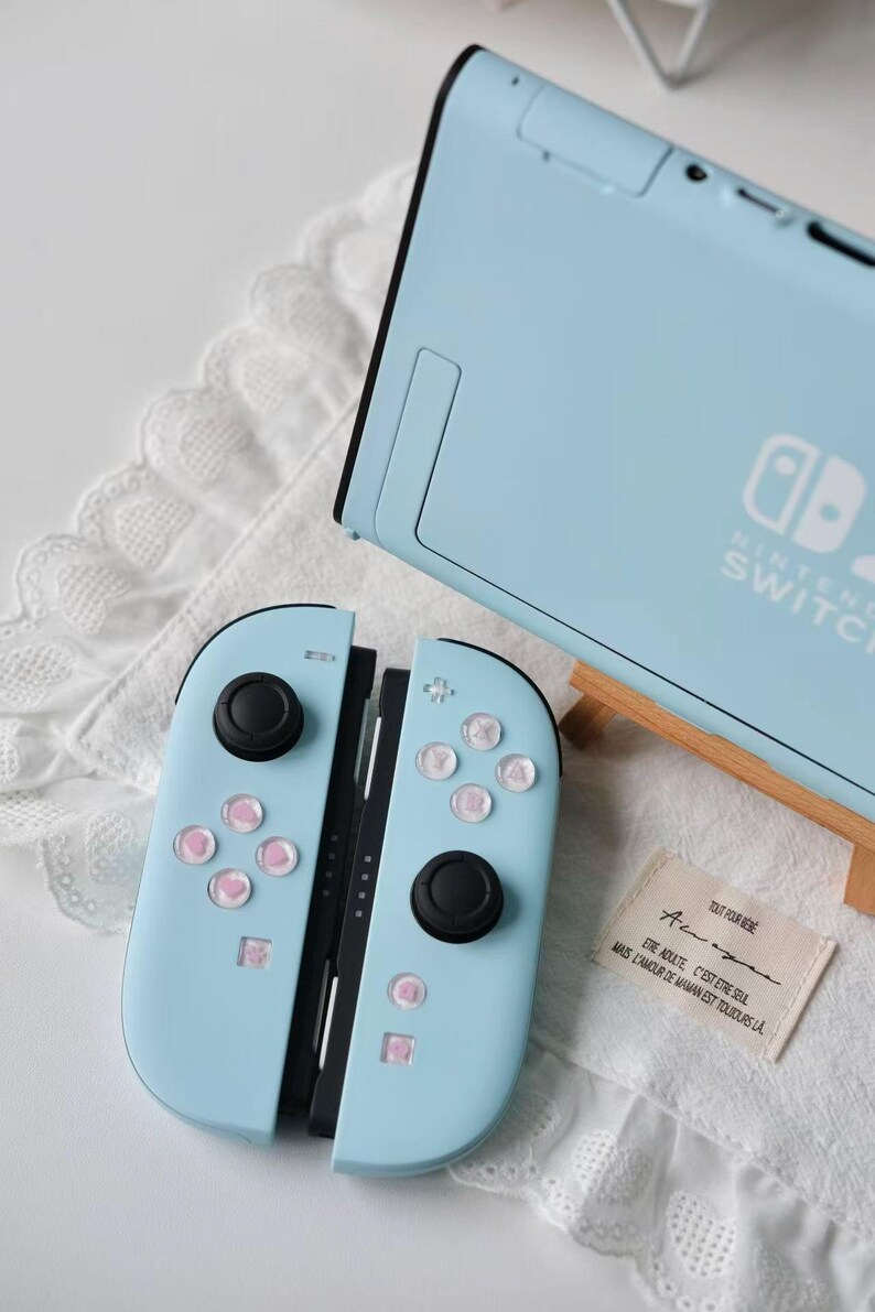 May include: A light blue Nintendo Switch console and a pair of matching Joy-Con controllers with black joysticks and light pink button accents. A small rectangular fabric tag with French text is placed next to the controllers on a white surface.