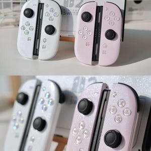 May include: Two sets of Nintendo Switch Joy-Con controllers. One set is white with iridescent buttons, the other is pink. Both sets have black control sticks and a glittery finish. A Nintendo Switch console is in the background.