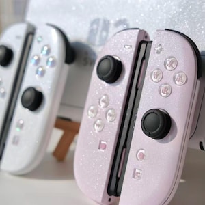May include: Two Nintendo Switch Joy-Con controllers, one white and one light pink, both with a glittery finish. Each controller has black joysticks and buttons. The pink controller is attached to a black device.