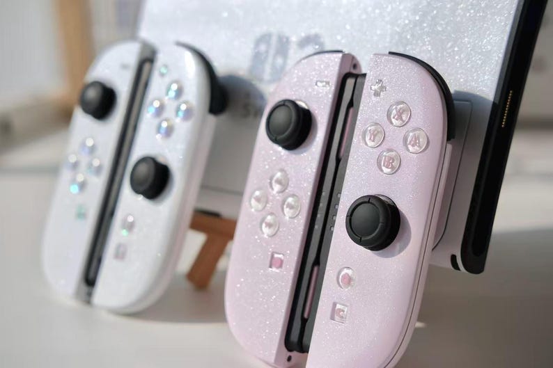 May include: Two Nintendo Switch Joy-Con controllers, one white and one light pink, both with a glittery finish. Each controller has black joysticks and buttons. The pink controller is attached to a black device.