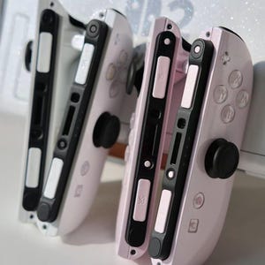 May include: Two pink and white game controllers with black buttons and joysticks. These controllers are designed for a gaming console. The background is white with a glittery effect.