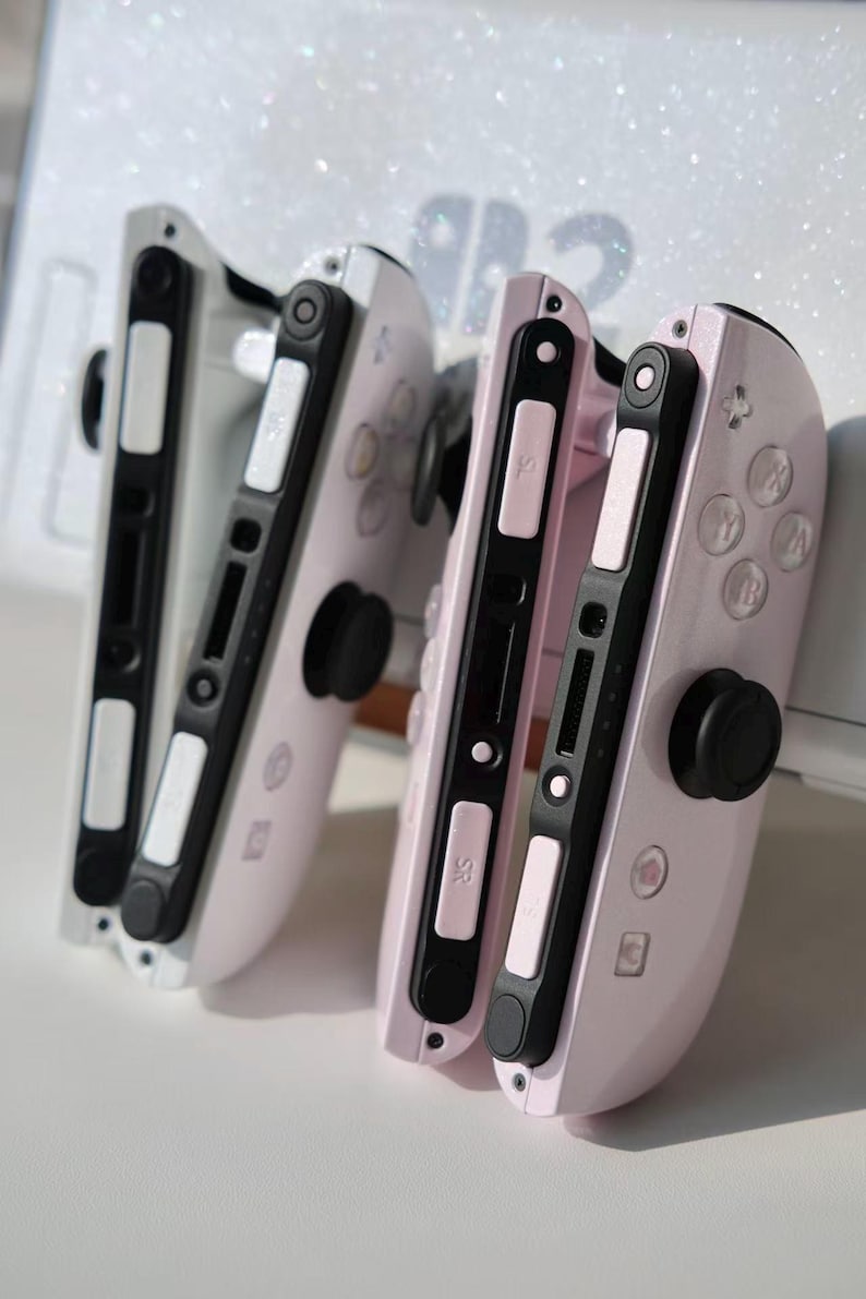 May include: Two pink and white game controllers with black buttons and joysticks. These controllers are designed for a gaming console. The background is white with a glittery effect.
