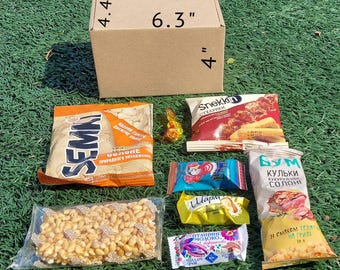 Può includere: Una scatola di cartone che misura 16 cm per 10 cm per 11 cm, piena di vari snack, tra cui cracker, cereali, caramelle e un sacchetto di semi di girasole. La scatola è appoggiata su un letto di erba verde.