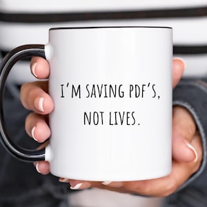 May include: White ceramic mug with a black handle and black rim. The mug has the text "I'm saving PDF's, not lives." printed in black.