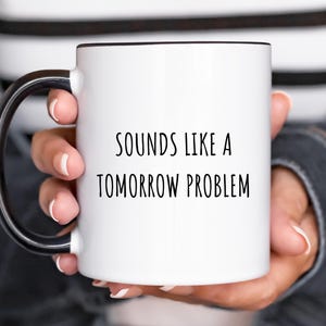 Könnte beinhalten: Weiße Keramik-Kaffeetasse mit schwarzem Henkel und schwarzem Rand. Die Tasse trägt den Text "Sounds like a tomorrow problem" in schwarzer Schrift.