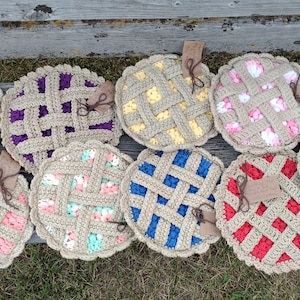 May include: A collection of ten handmade crochet pie-shaped items. Each pie features a tan lattice crust and a colorful filling, including blue, purple, pink, yellow, and red. Each pie has a small brown tag attached. The items are arranged on a wooden bench.