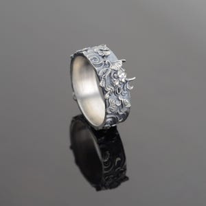 May include: A silver ring with a detailed floral design. The ring is wide and has a textured surface.