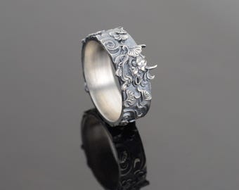 May include: A silver ring with a detailed floral design. The ring is wide and has a textured surface.