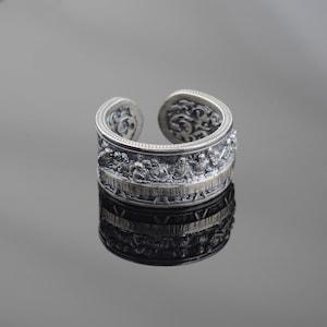 Könnte beinhalten: Ein silberner Ring mit einem detaillierten Design einer Szene mit mehreren Figuren. Der Ring ist an einer Seite offen und hat eine strukturierte Oberfläche.
