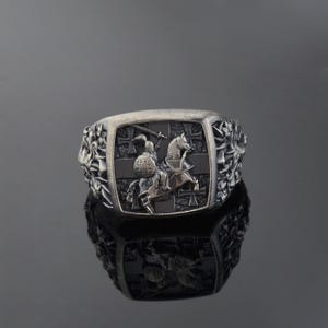 Könnte beinhalten: Ein silberner Ring mit einer schwarzen Emaille-Einlage, die einen Ritter auf einem Pferd mit einem Schwert darstellt. Der Ritter trägt eine Rüstung und einen Helm. Der Ring ist von einem Rand aus Kreuzen umgeben.