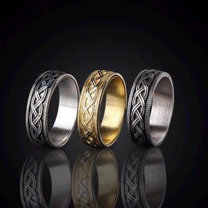 May include: Three rings with Celtic knot designs. One ring is gold-colored, while the other two are silver-colored. The rings are displayed on a reflective black surface, showcasing their intricate braided patterns.