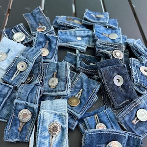 May include: A collection of denim jean button extenders in various shades of blue. Each extender is made from a small piece of denim and features a metal button and buttonhole. Some buttons have text.