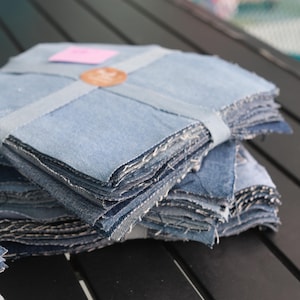 May include: Stacks of light blue denim fabric squares, neatly bundled and secured with a matching denim ribbon. A small pink tag and a brown circular tag are visible on top. The edges of the fabric are frayed.