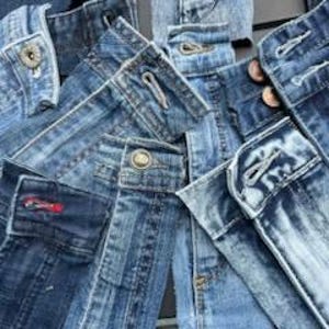 May include: Close-up of several pairs of blue denim jeans, showcasing waistbands, belt loops, and button closures. The jeans vary in shades of blue, from light wash to darker indigo, with visible stitching and metal hardware.