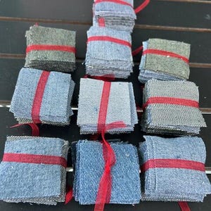 May include: Nine stacks of small, square denim fabric pieces in various shades of blue and gray. Each stack is tied with a bright red ribbon. The arrangement suggests a craft or DIY project, possibly for quilting or other textile arts.