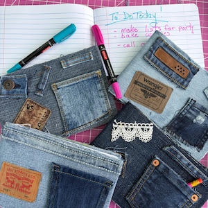 May include: Three denim pouches crafted from repurposed jeans, featuring various pockets, leather brand labels, and a white lace trim. A notebook with the handwritten text To Do Today and two permanent markers in blue and pink are positioned nearby.