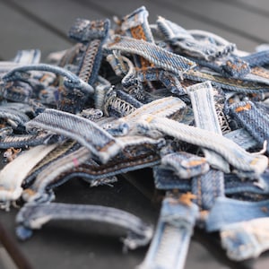 May include: A pile of denim fabric scraps in various shapes and sizes. The scraps are primarily blue with visible stitching and frayed edges, suggesting they are from jeans or other denim garments. The image is a close-up, highlighting the texture and details.