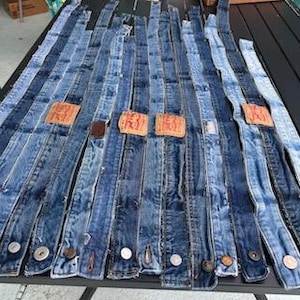 May include: A collection of denim belts in various shades of blue, arranged on a dark surface. Each belt features a different wash and texture, with visible stitching, metal buttons, and leather patches. The belts are laid out in a parallel fashion.