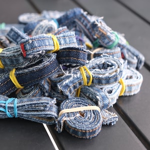 May include: A pile of rolled denim strips, secured with colorful rubber bands. The blue jean material shows visible stitching and varying shades of blue. The strips are tightly wound, creating a textured and organized appearance.