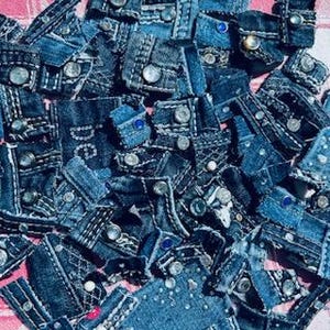 May include: A pile of small, irregularly shaped denim patches with silver-colored buttons and white stitching. Some patches have small blue gemstones. The denim is various shades of blue.