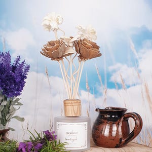 May include: A clear glass diffuser bottle with a wooden lid and brown wooden sticks. The bottle has a label that says "Dawn Lit" and "Countryside". The diffuser is surrounded by dried flowers and lavender.