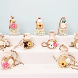 May include: A selection of car air fresheners in glass bottles. Each bottle contains a light-coloured liquid and is decorated with a wooden cap and a decorative element, such as a daisy, teddy bear, or flower. The bottles are displayed on a white surface.
