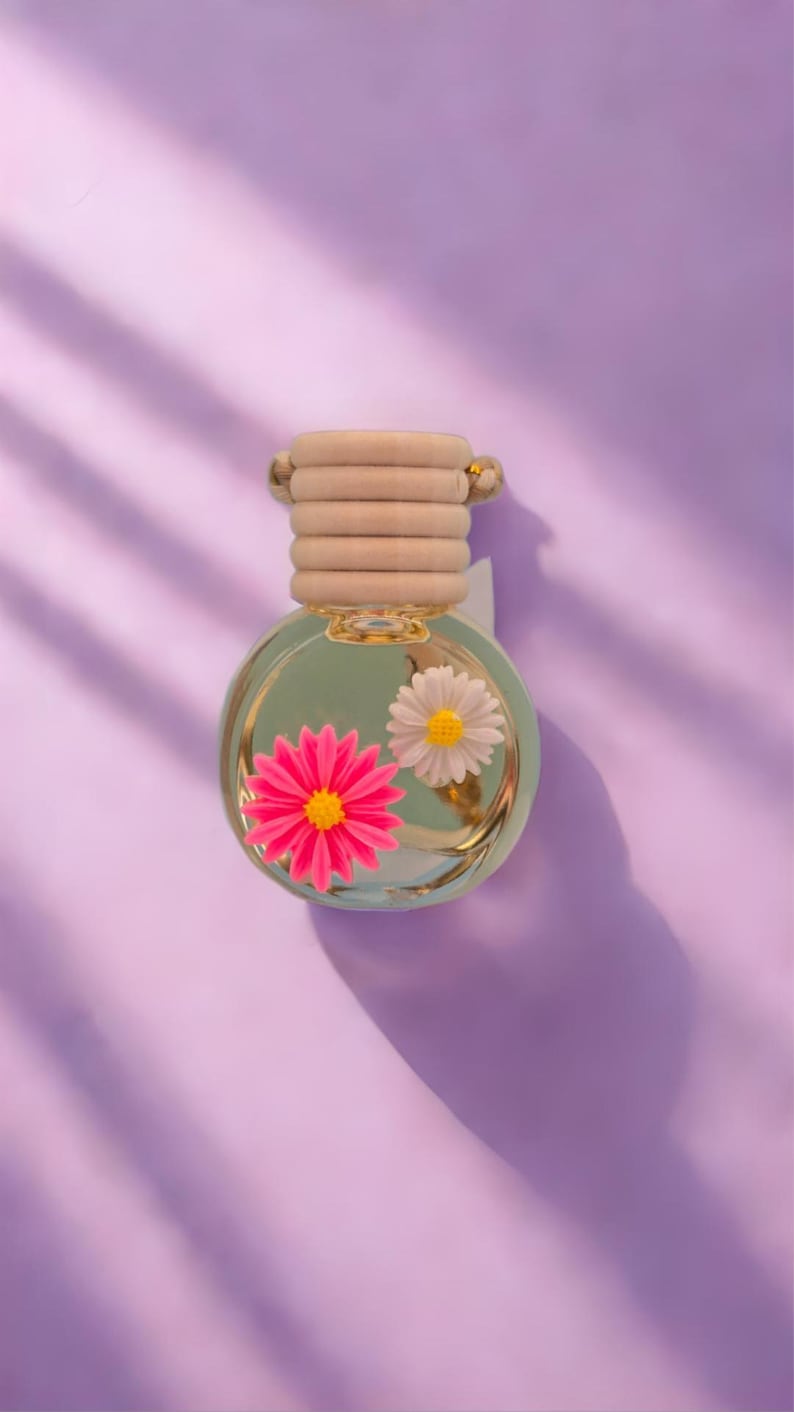 May include: A clear glass bottle with a gold lid and a beige fabric ring. The bottle contains a clear liquid and two small plastic flowers, one pink and one white.