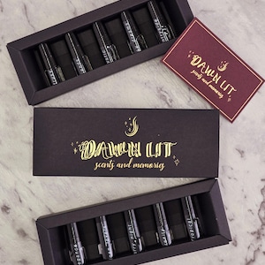 May include: Three black gift boxes with gold text reading "DAWN LIT scents and memories" are shown. Each box contains perfume vials labelled with names such as "Elegance" and "Tropical".