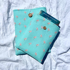 May include: Two turquoise book sleeves with a pink flamingo pattern. Each sleeve has a wooden button closure. One sleeve holds a book with the text "The New York Times Bestseller" visible.
