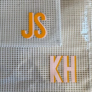 May include: A close-up of a white mesh fabric with two sets of initials in orange and pink. The initials "JS" are in orange, and the initials "KH" are in pink with an orange outline. The fabric is layered.