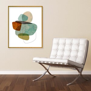 May include: Abstract art print with a gold frame, featuring a composition of overlapping shapes in shades of teal, brown, and yellow, displayed on a wall above a white leather armchair with a chrome base.