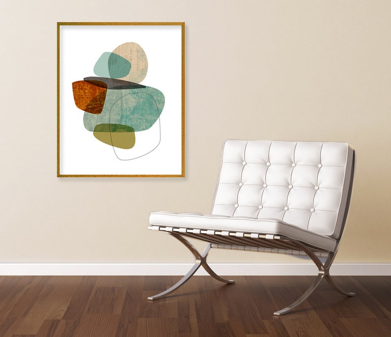May include: Abstract art print with a gold frame, featuring a composition of overlapping shapes in shades of teal, brown, and yellow, displayed on a wall above a white leather armchair with a chrome base.
