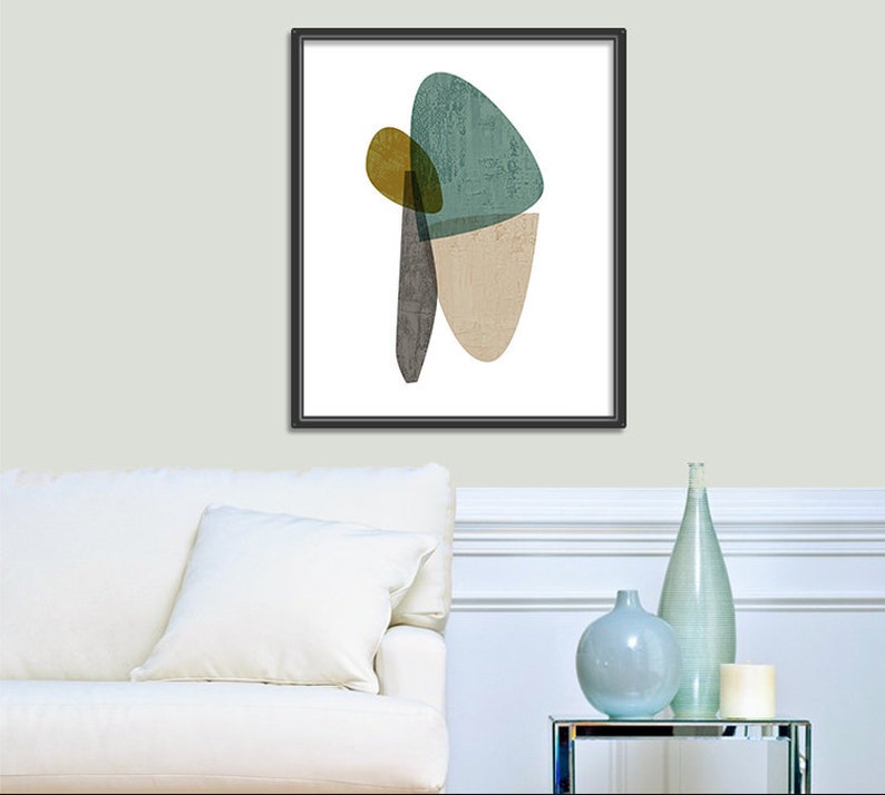 Puede incluir: Impresi&oacute;n de arte abstracto con formas superpuestas en tonos de verde azulado, marr&oacute;n, beige y amarillo. La impresi&oacute;n est&aacute; enmarcada en negro y se muestra en una pared blanca sobre un sof&aacute; blanco.