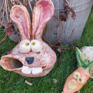 Könnte beinhalten: Ein skurriler Keramik-Hasenkopf mit großen Ohren, großen Augen und einem grinsenden Mund. Der Hase ist hellbraun mit rosa Akzenten in den Ohren. Daneben eine Keramikkarotte mit grünen Blättern. Die Objekte liegen auf grünem Gras.