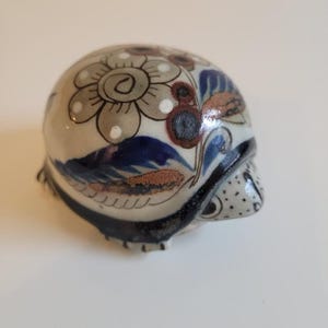 May include: A ceramic skull figurine with a floral design. The skull is painted with a cream base colour and features a large grey flower with white dots, blue leaves, and brown accents. The edges are outlined in black.