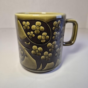 May include: A ceramic mug featuring a dark brown and olive green glaze with a raised, embossed floral pattern. The design includes stylised four-petaled flowers and a curved, abstract shape, complemented by a sturdy handle on the right side.