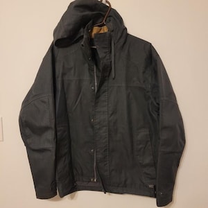 May include: A dark grey hooded jacket with a zip and snap fastenings. The jacket features a hood, pockets, and a brown lining. It is hanging on a brown hanger.