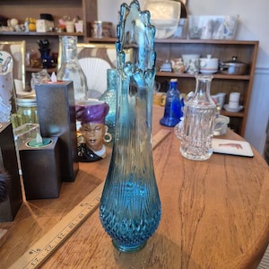 May include: A tall, turquoise glass vase with a textured, diamond-patterned base and a flared, scalloped top. The vase is approximately 30 cm tall and stands on a wooden table, showcasing its elegant design.