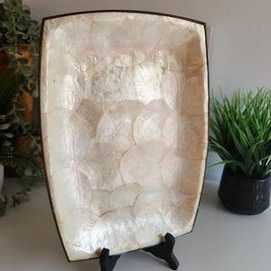 Peut inclure: Plateau décoratif rectangulaire avec incrustation de nacre. Le plateau a un cadre en métal marron foncé et est soutenu par un support noir. La surface est constituée de pièces irisées en forme de coquillage. Le plateau est vide.