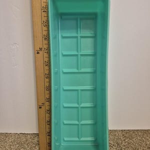 May include: A rectangular, light teal plastic mould with a grid-like pattern. A wooden ruler with inch markings is positioned next to the mould. The ruler shows measurements from 61 to 89 cm.